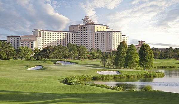 The exterior of Rosen Shingle Creek, a large hotel surrounded by rolling golf course greens, a lake, and trees.